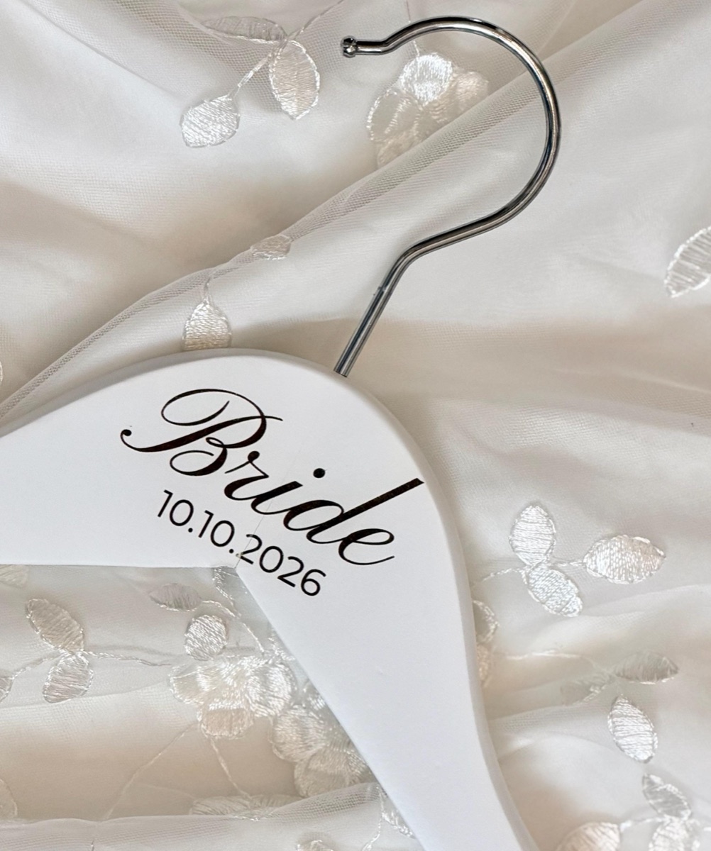 Close-up of a white wooden hanger engraved Bride 10.10.2026 in a script font